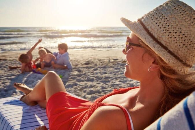 A woman wearing a hat and sunglasses relaxes on a beach lounger while her family enjoys the sand in the background, making it easier to remember essential items when utilizing smart packing hacks.